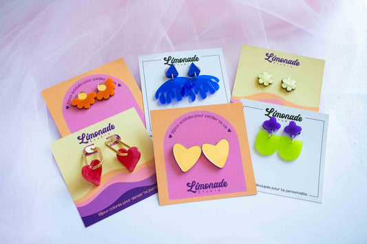 Assortiment de boucles d'oreilles acryliques Limonade Studio en formes variées — fleurs orange, cœurs dorés, vagues bleues et étoiles vertes sur cartes colorées