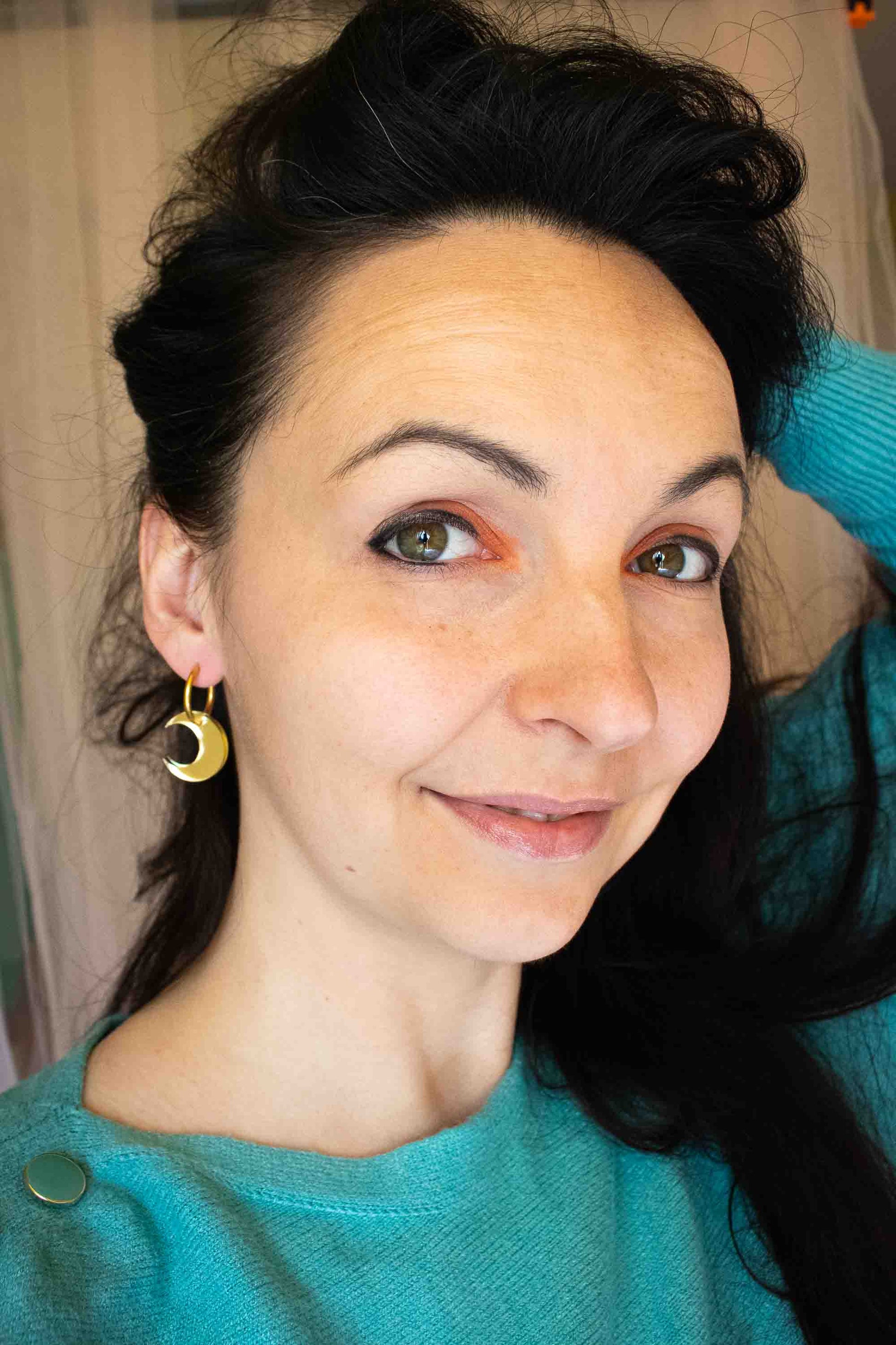 Portrait lifestyle de la créatrice portant les boucles huggies lune dorée miroir, bijou fait main Limonade Studio, pull turquoise, ambiance chaleureuse