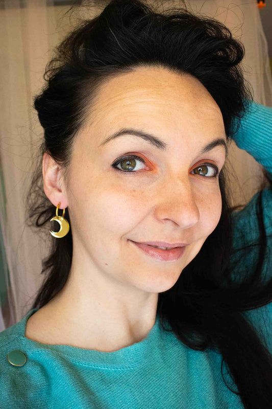 Portrait lifestyle de la créatrice portant les boucles huggies lune dorée miroir, bijou fait main Limonade Studio, pull turquoise, ambiance chaleureuse