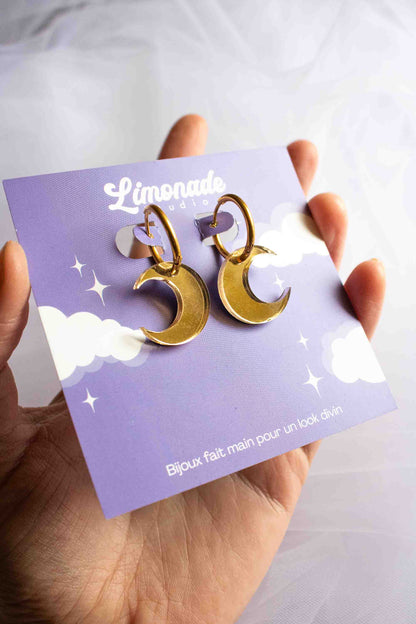 Boucles d'oreilles huggies dorées avec pendentif lune en acrylique miroir doré, faites main par Limonade Studio, sur cartonnette violette