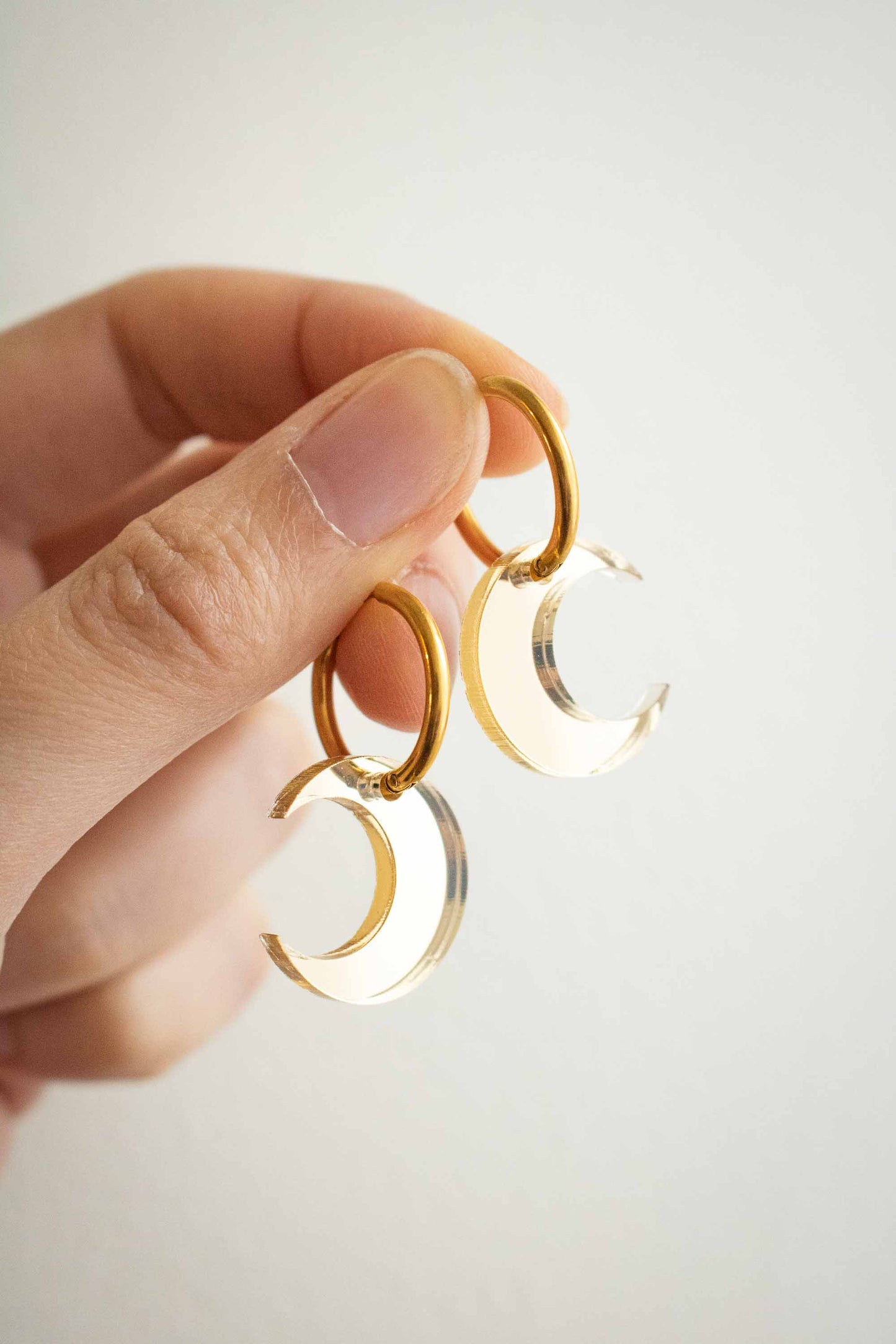 Boucles d'oreilles huggies dorées avec pendentif lune en acrylique transparent miroir, bijou fantaisie fait main Limonade Studio, tenues entre les doigts