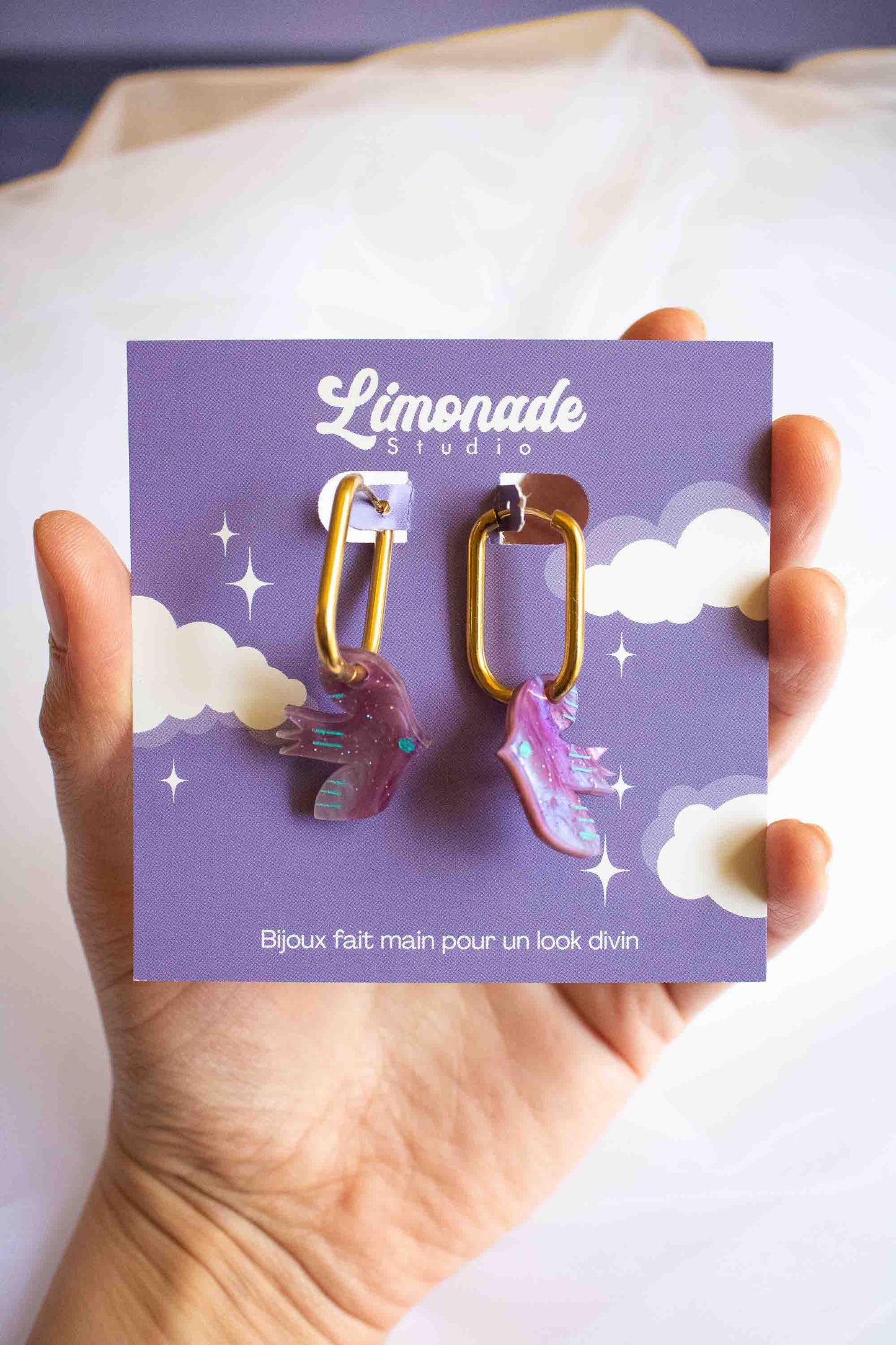 Boucles d'oreilles huggies oiseau violet en acrylique marbré, anneau acier doré, fait main par Limonade Studio, sur cartonnette tenue en main