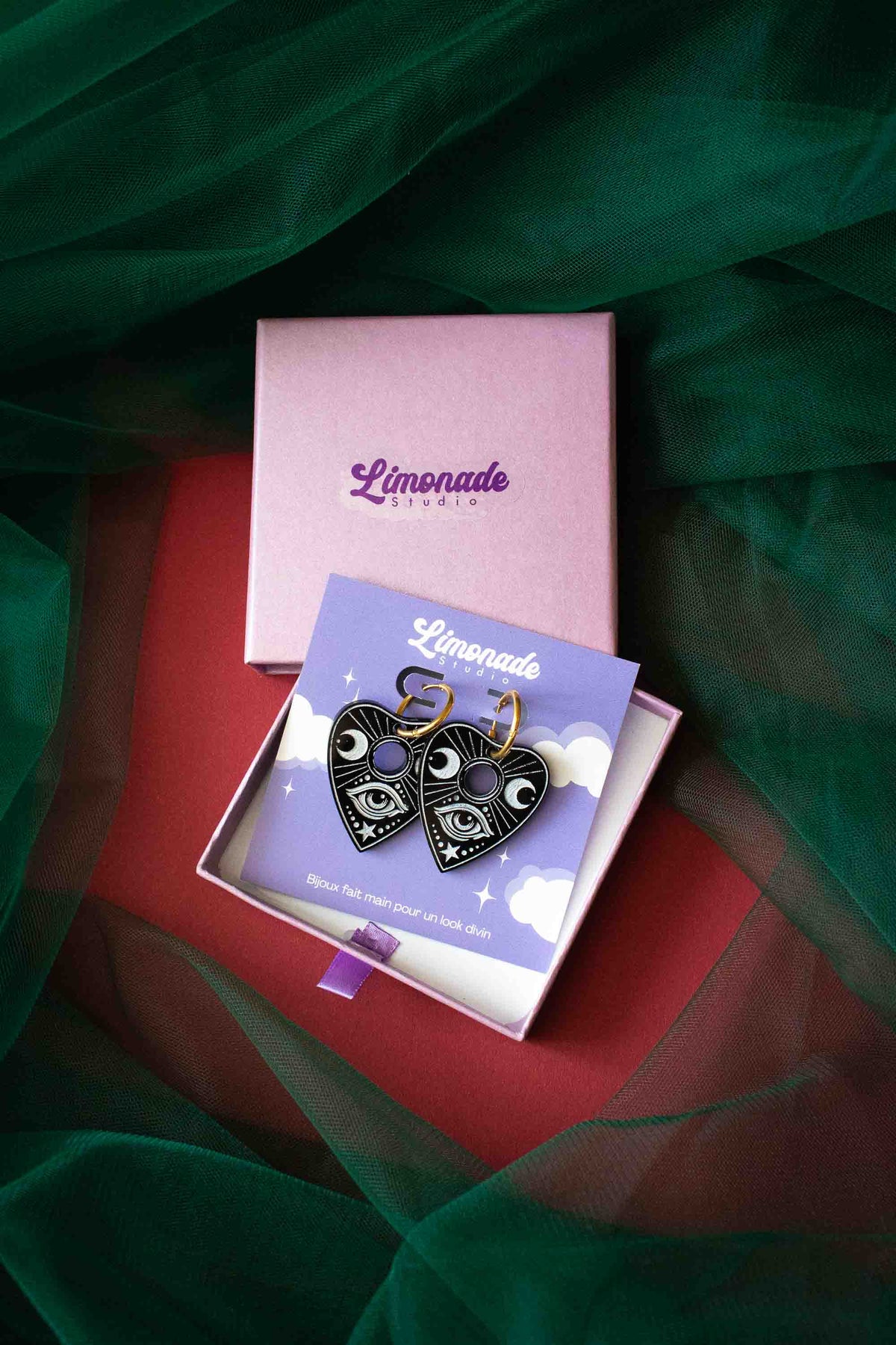 Paire de boucles Ouija présentée dans un coffret violet ouvert sur un tissu rouge et vert