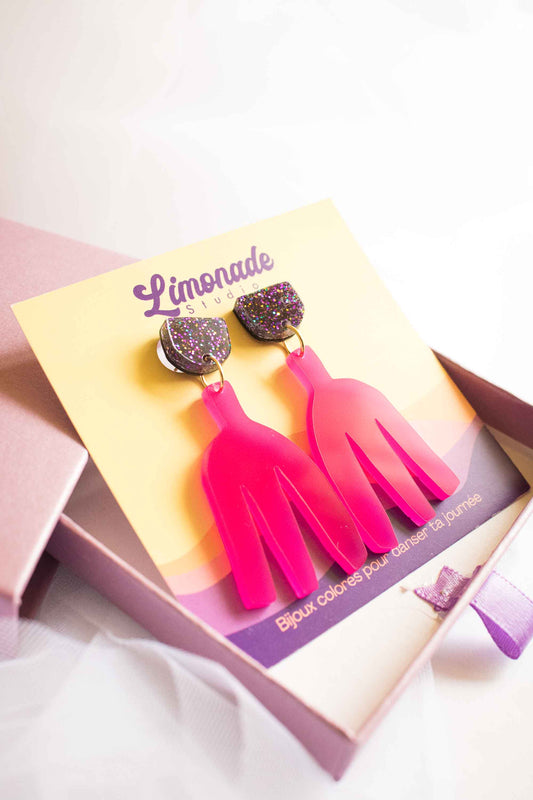 Boîte cadeau rose ouverte avec les boucles pendantes fuchsia sur cartonnette Limonade Studio, ruban violet, vue de côté
