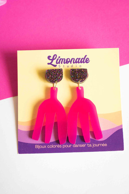 Paire de boucles pendantes fuchsia clou pailleté sur cartonnette Limonade Studio, fond bicolore rose et blanc