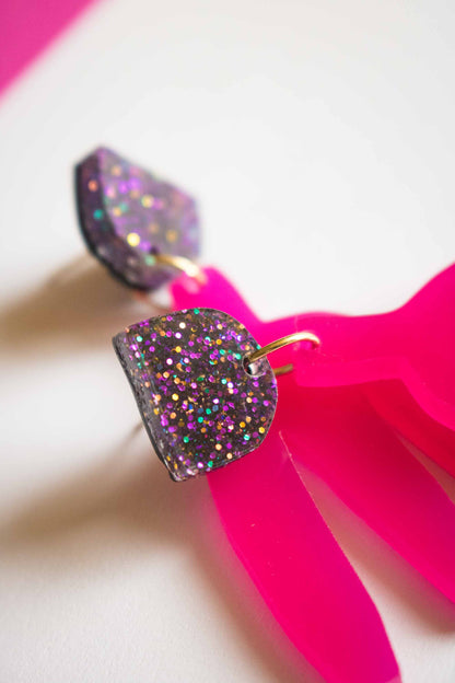 Gros plan sur une boucle pendante fuchsia acrylique laser, détail du clou pailleté holographique et de l'anneau doré de liaison