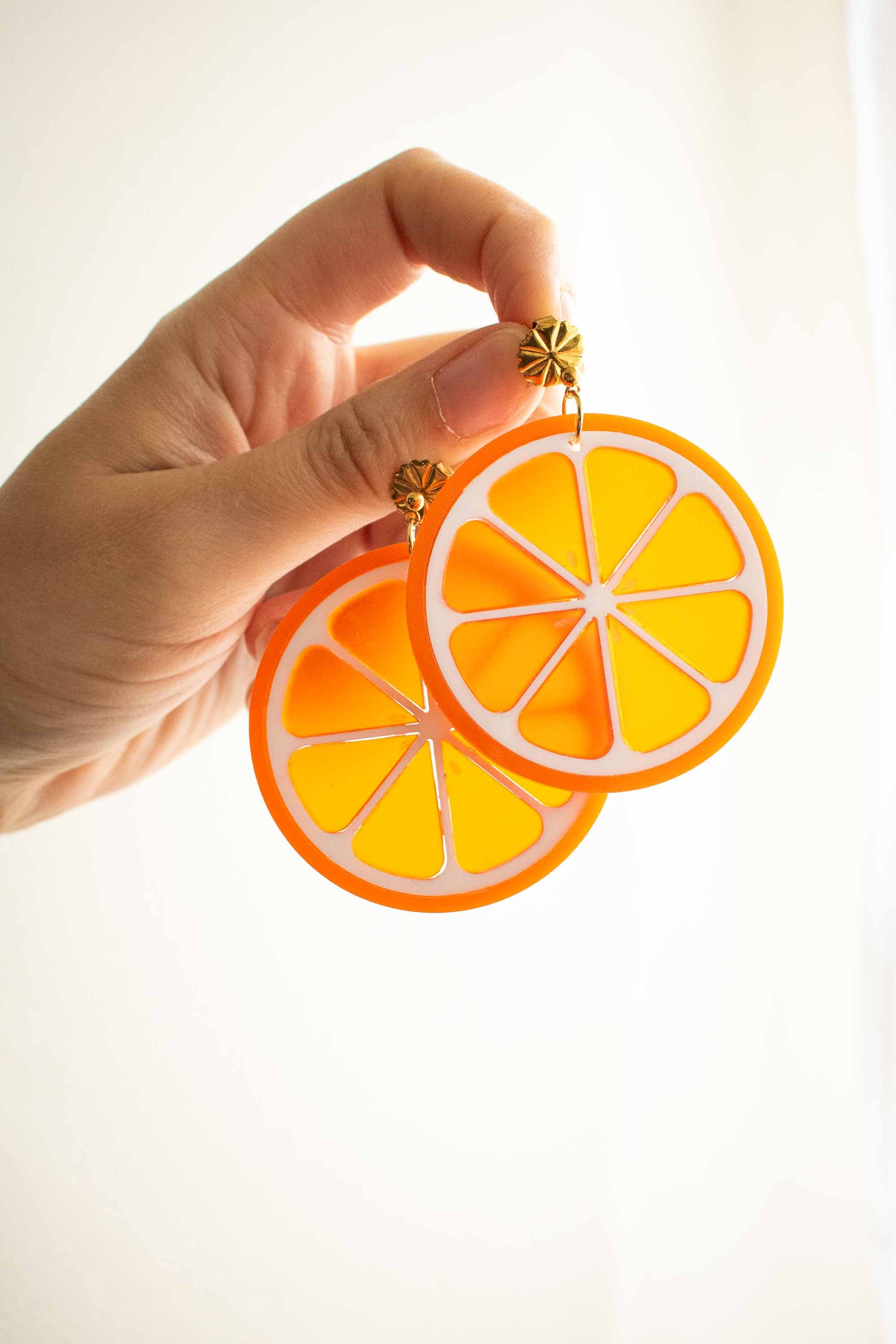 Boucles d'oreilles pendantes orange acidulé en acrylique fait main, bijou fantaisie puce dorée, Limonade Studio, tenues entre les doigts sur fond blanc