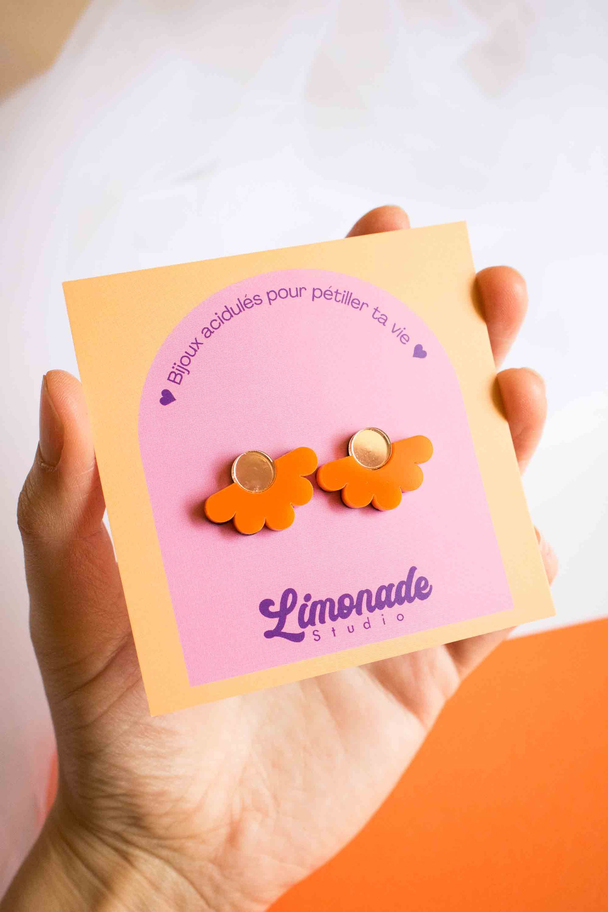 Main tenant la cartonnette Limonade Studio avec les puces fleur orange en acrylique découpé au laser, fond bicolore