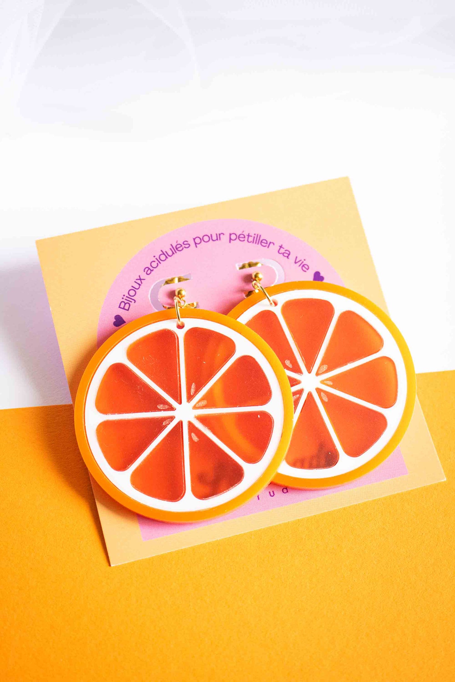 Boucles d'oreilles pendantes tranche d'orange en acrylique fait main, cadeau femme, puce dorée, Limonade Studio, présentées sur cartonnette rose et jaune