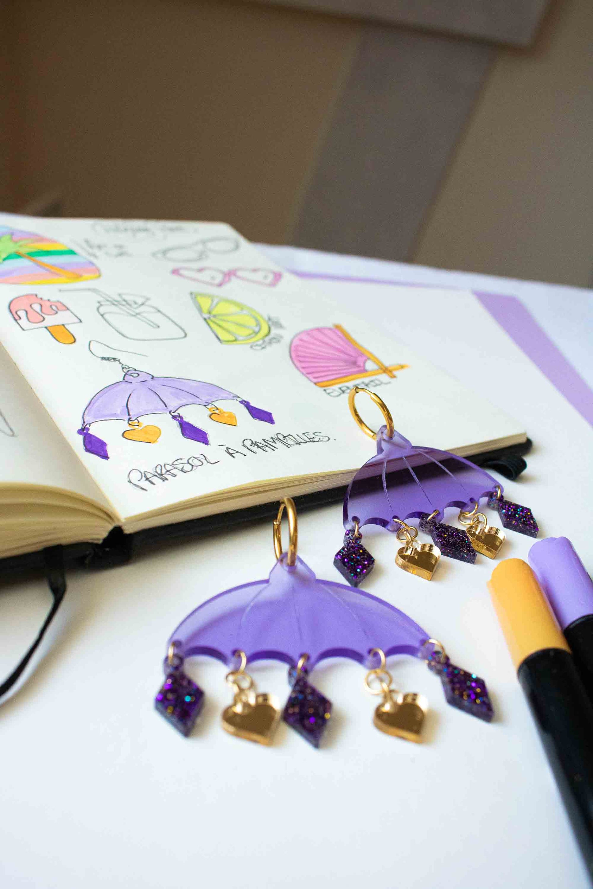 Boucles d'oreilles violettes en acrylique decoupe laser, bijou colore artisanal Limonade Studio, presentees dans un atelier avec carnet de croquis.