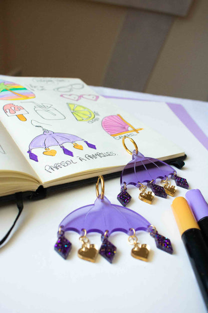 Boucles d'oreilles violettes en acrylique decoupe laser, bijou colore artisanal Limonade Studio, presentees dans un atelier avec carnet de croquis.
