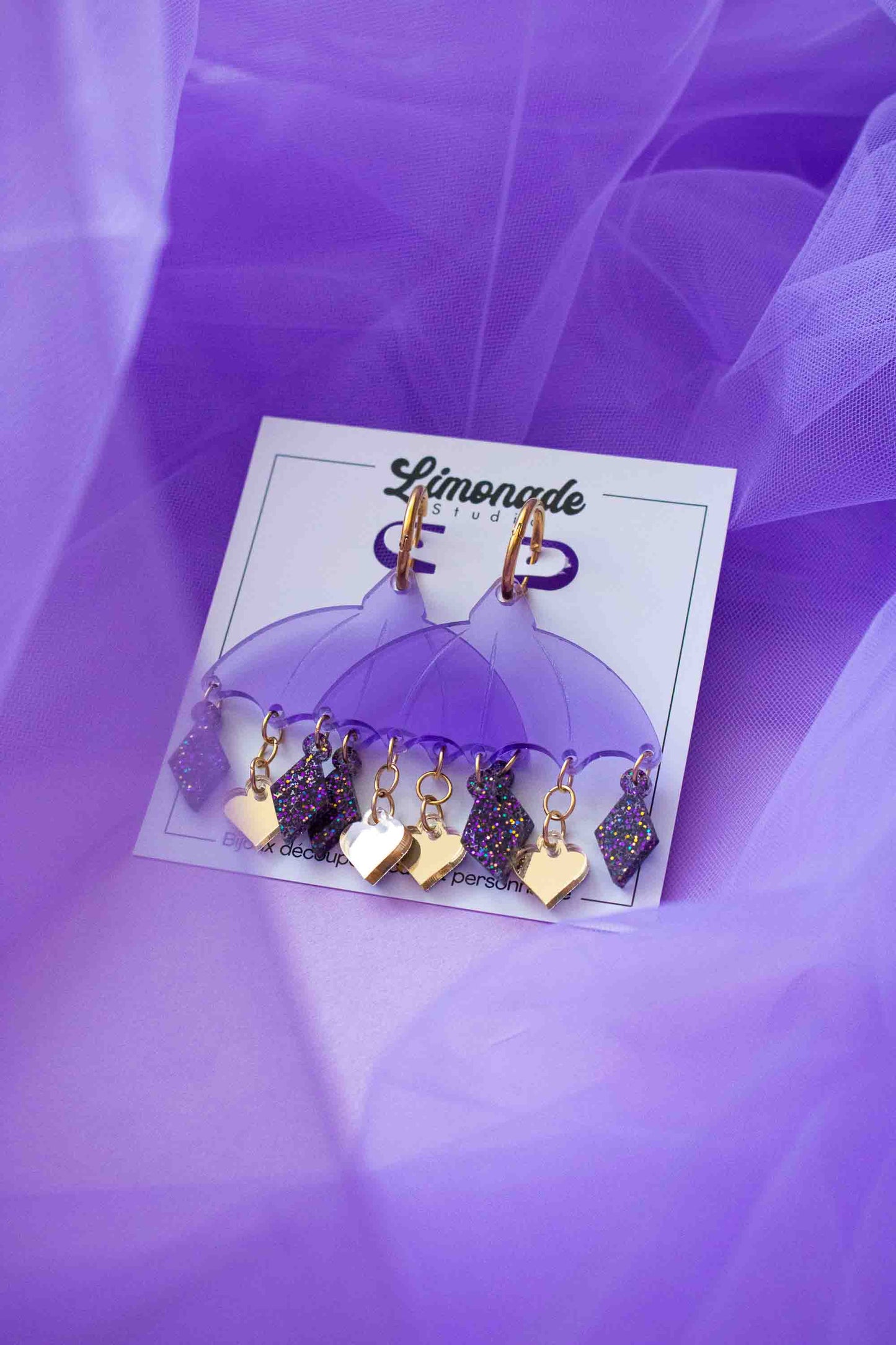 Boucles d'oreilles violettes en acrylique decoupe laser, bijou colore artisanal Limonade Studio, presentes sur cartonnette avec charms pendants.