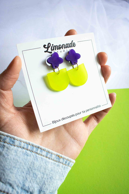 Boucles pendantes fleur violette et vert acrylique dépoli fait main Limonade Studio, sur cartonnette tenue en main, fond vert et blanc