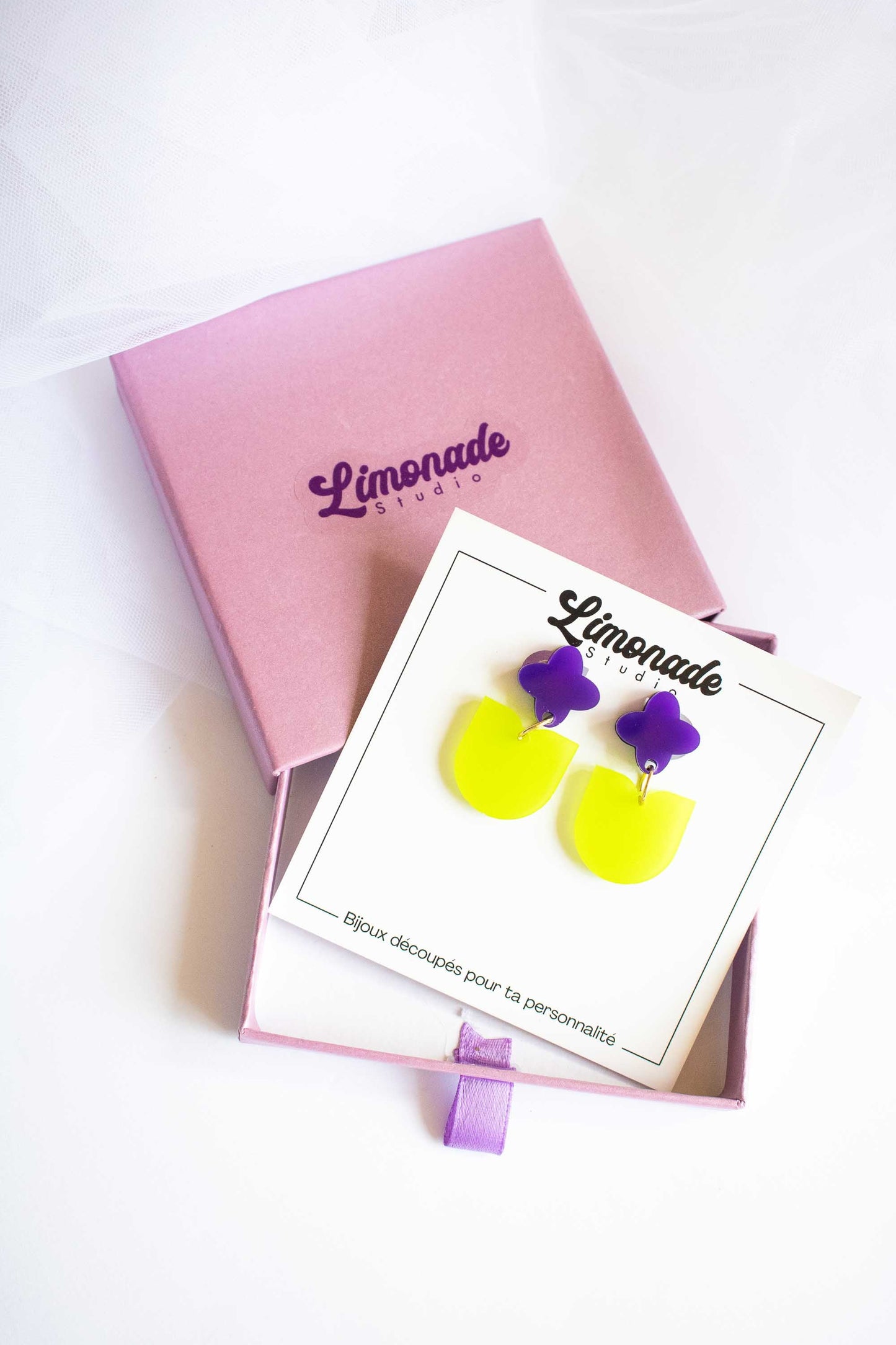 Boucles d'oreilles fantaisie fleur violette vert acrylique fait main Limonade Studio, sur cartonnette posée sur boîte cadeau violette