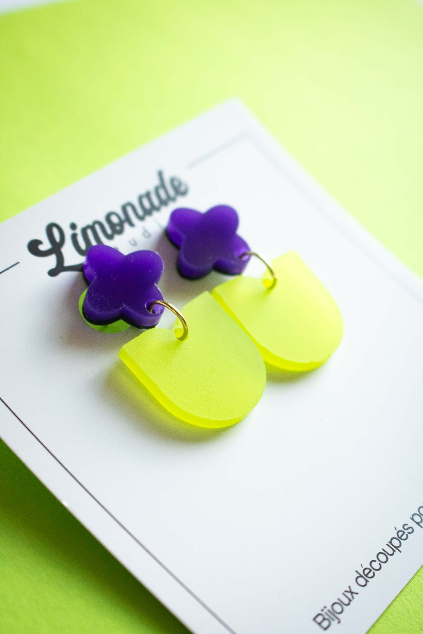 Paire de boucles pendantes fleur violette et vert acrylique dépoli fait main, sur cartonnette Limonade Studio, fond vert citron